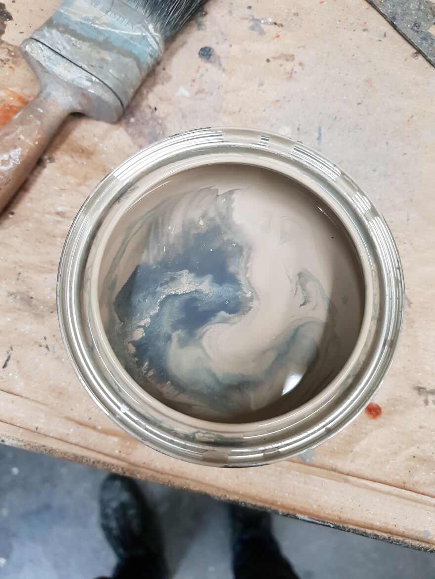 Swirled paint in a can resembling accidental art on a wooden surface next to a brush.