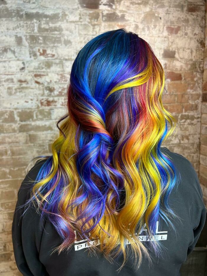 Vibrant rainbow hair color transformation with shades of blue, yellow, and red against a brick wall background.