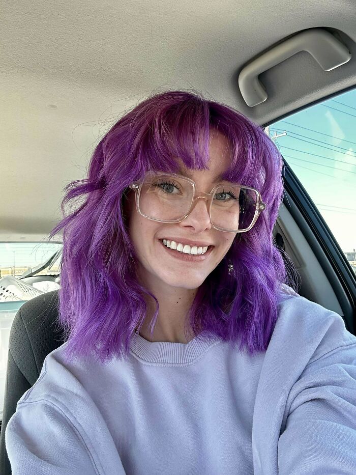 Person in a car with purple hair, wearing glasses and a light sweater, smiling. Changing hair color to vibrant shades.