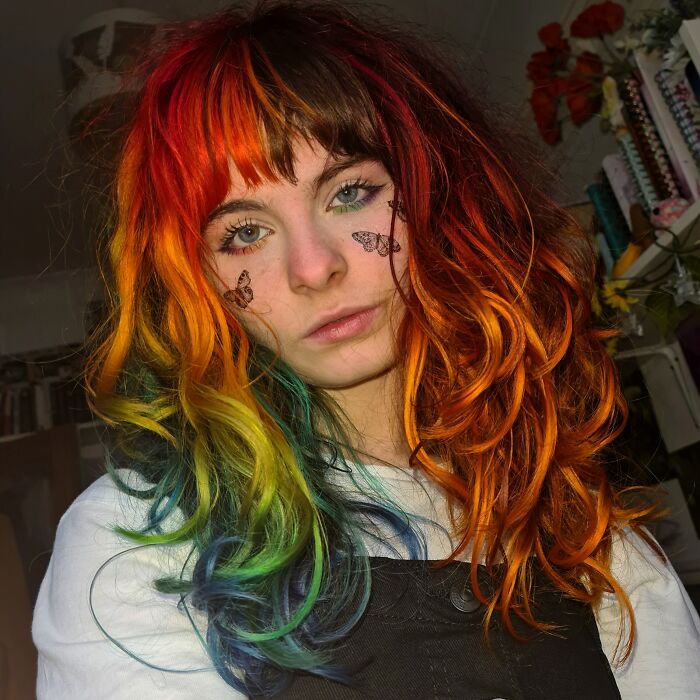 Vibrantly changing hair color with rainbow hues, worn by a person with butterfly face tattoos, in a casual setting.