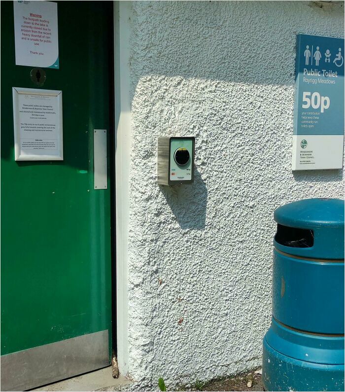 Public toilet in the UK with coin-operated entry, green door, and blue trash bin, showcasing British quirks and humor.