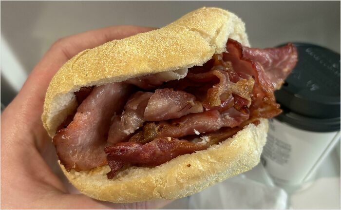 A hand holding a bacon sandwich with a coffee cup in the background, illustrating British humor.