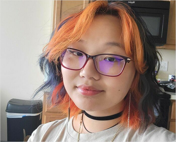 Person with changing-hair-color, featuring orange and dark tones, wearing glasses and a choker, indoors.