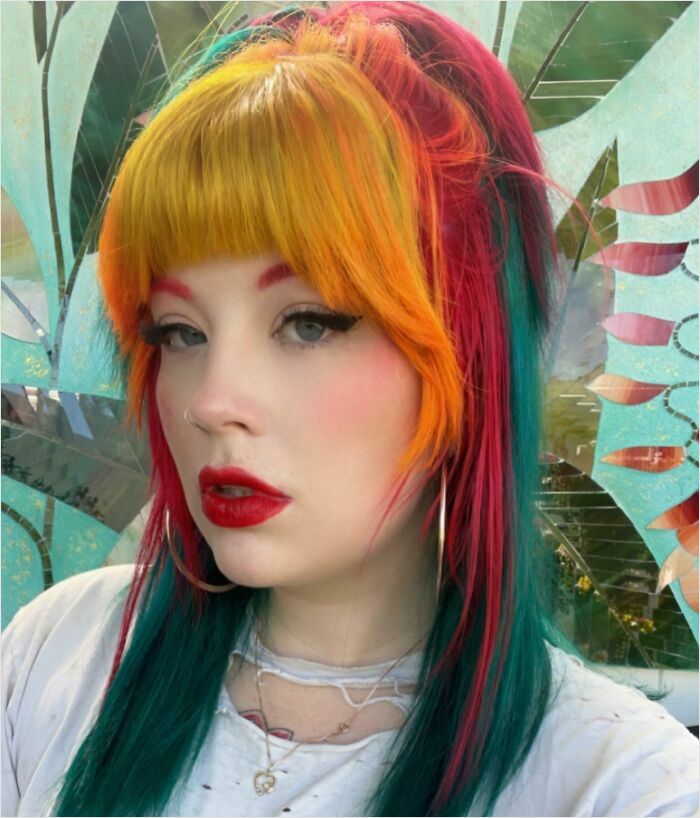 A person with vibrant, multi-colored hair featuring shades of yellow, red, and green against a decorative background.