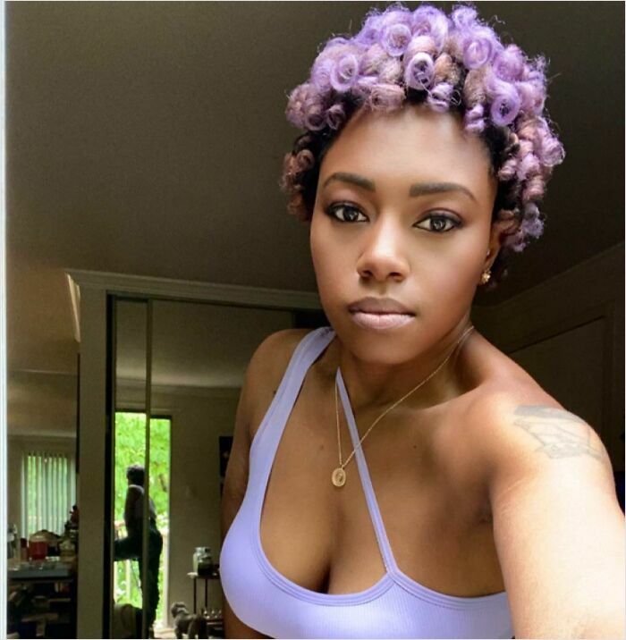 Woman with changing hair color, curly purple hairstyle, wearing a lavender top, posed in a sunlit room.