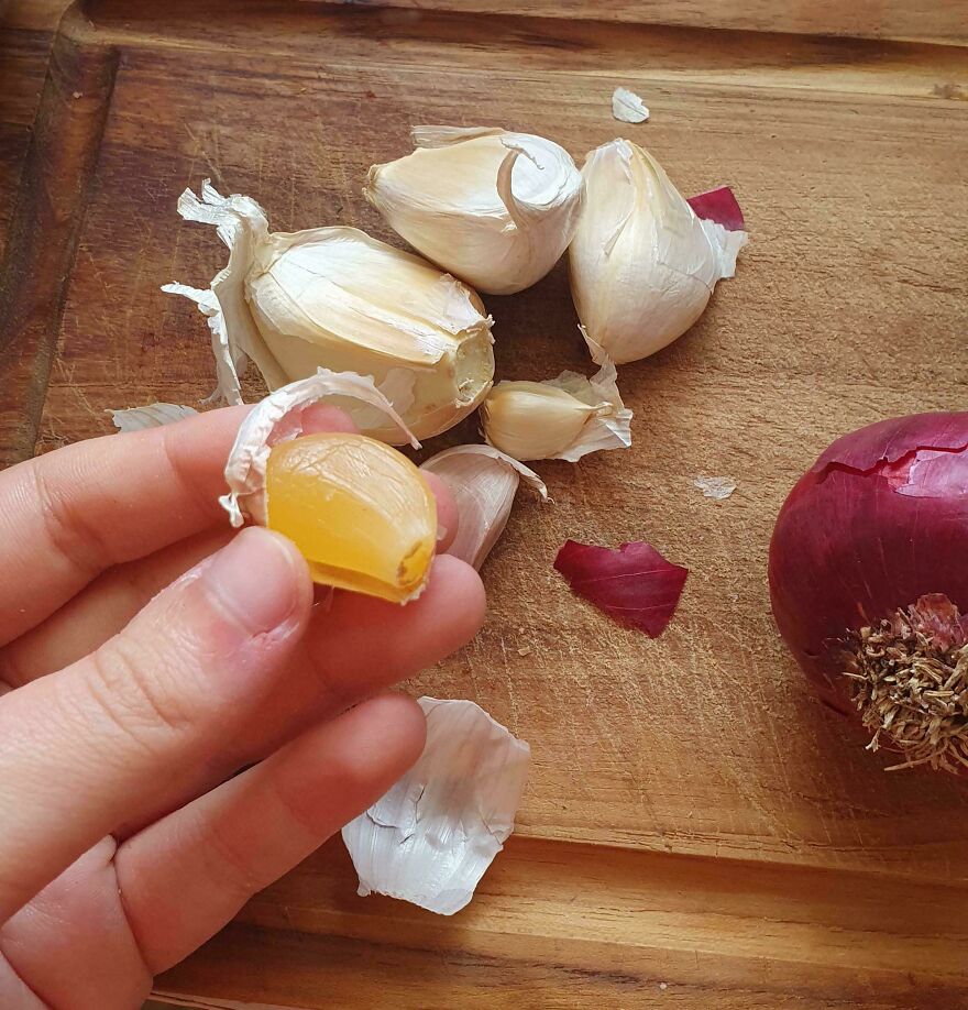 A hand holding a garlic clove slice that looks like an egg yolk, with more garlic and a red onion on a cutting board.