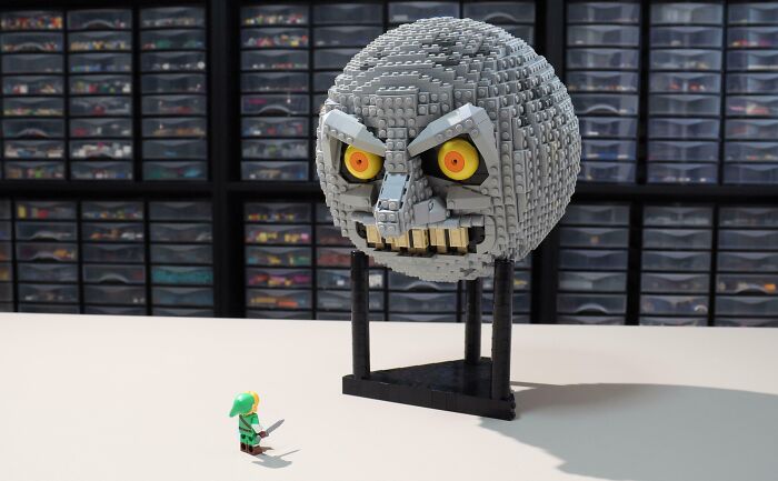 LEGO model of a menacing moon face with a small figure in green clothing on display in front of storage shelves.