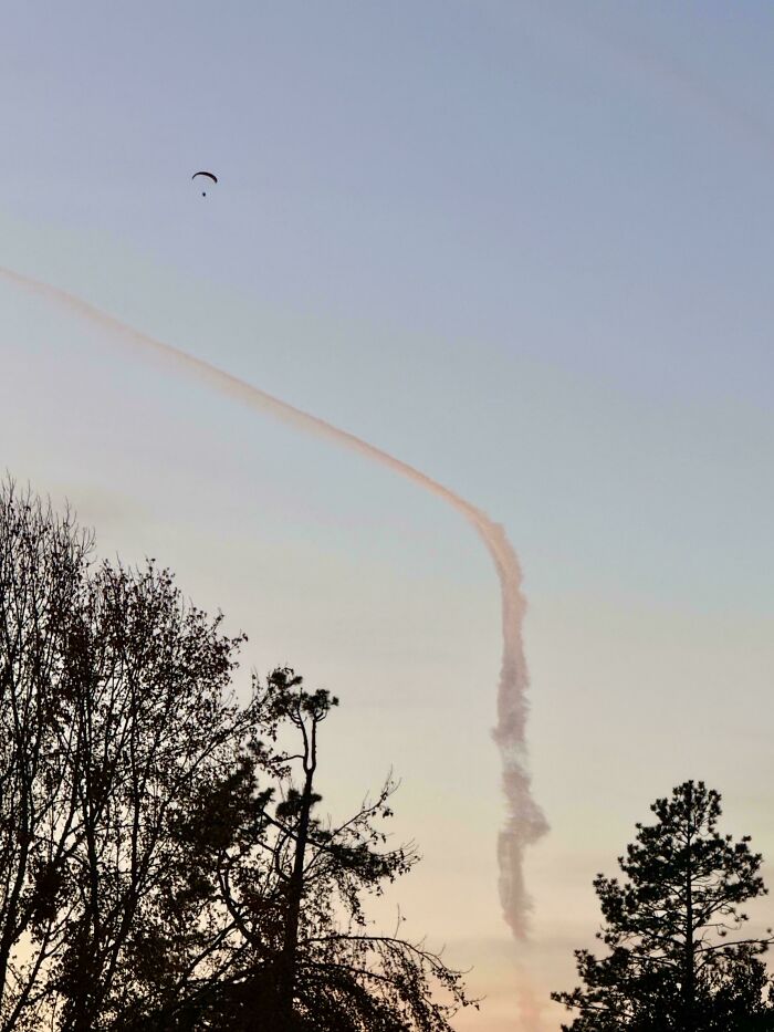 Paraglider above trees with a confusing perspective, creating an illusion of a smoke trail in the sky.