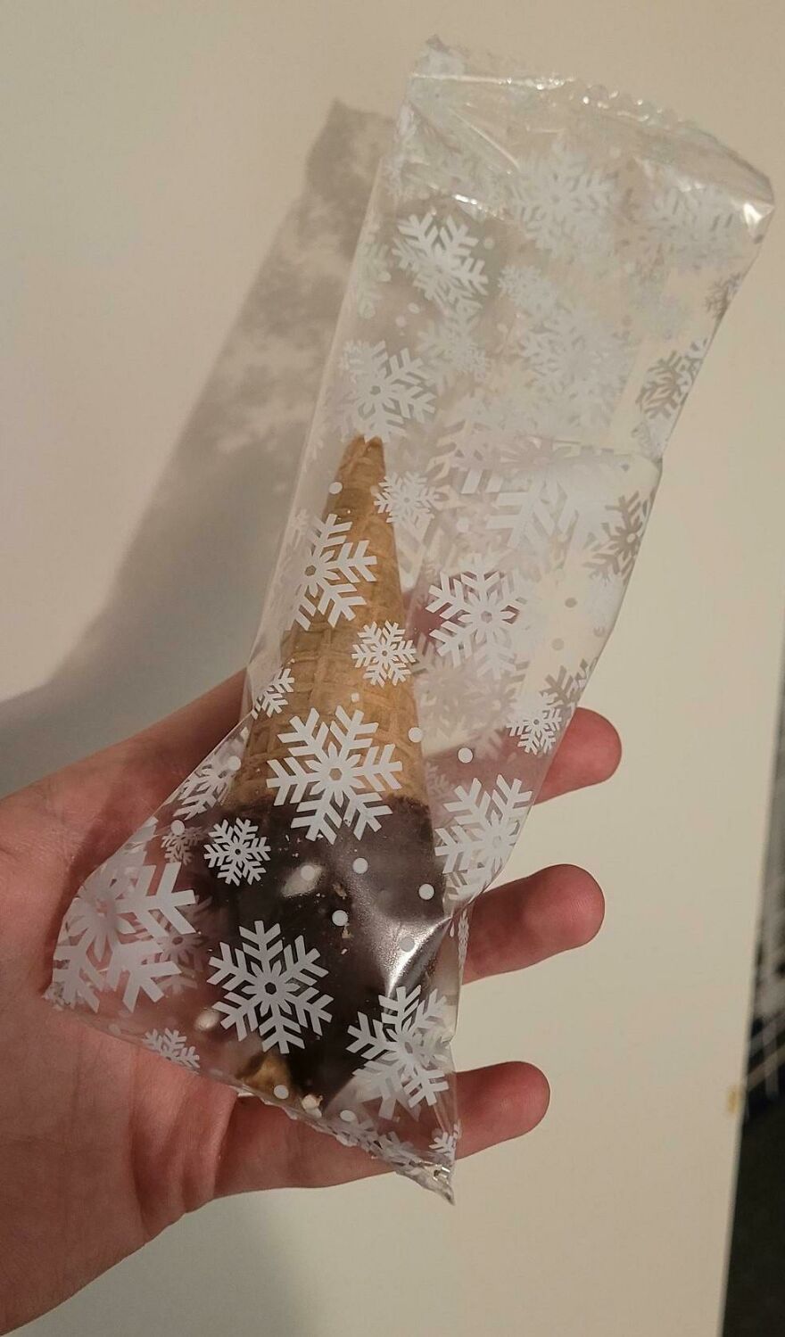 A hand holding a chocolate-dipped cone in decorative unnecessary packaging with snowflakes.