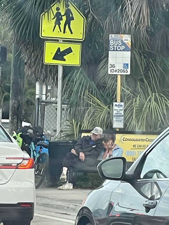 Confusing perspective at a bus stop with two people seated, surrounded by bicycles and cars.