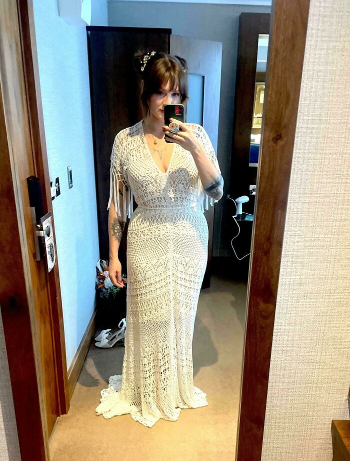 Person in mirror wearing intricately crocheted white dress in a room setting.