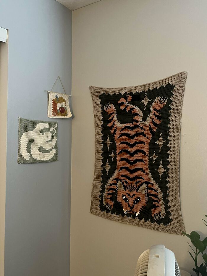 Crocheted wall art featuring a tiger and two abstract designs hanging on a beige wall.