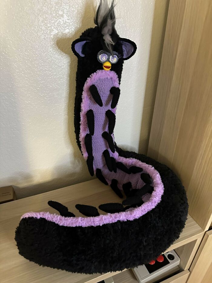 A creatively crocheted plush toy with a long, snake-like body, featuring purple and black colors, displayed on a shelf.