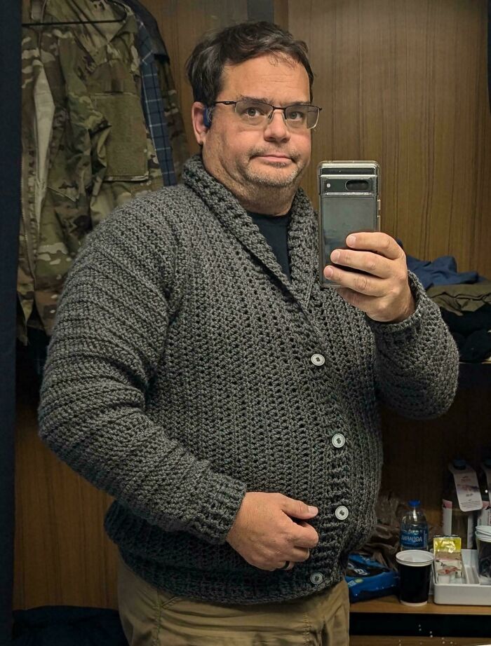 Man wearing a handmade crocheted gray cardigan, taking a mirror selfie in a room.