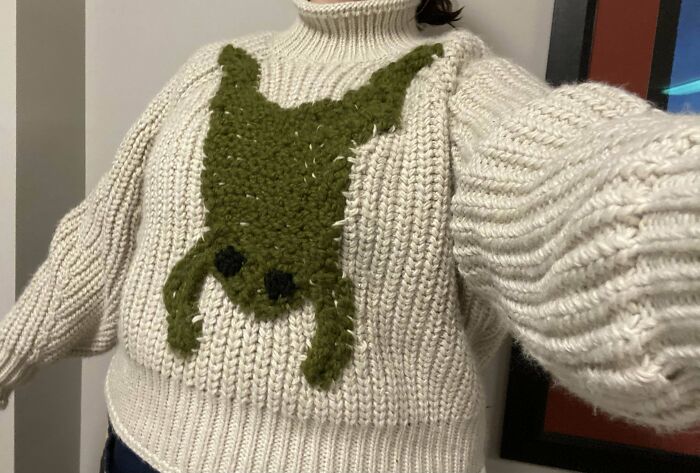 A person wearing a crocheted sweater with a green animal design on the front.