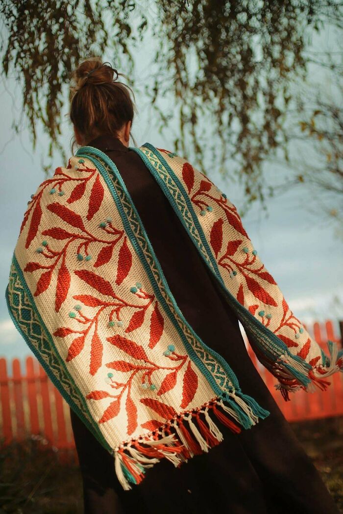 Person wearing a crocheted shawl with red leaf patterns and turquoise edges outdoors.