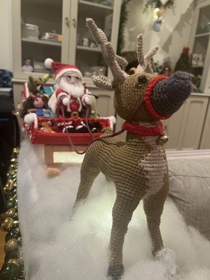 Crocheted reindeer pulling Santa in a sleigh, surrounded by fluffy snow in a festive setting.