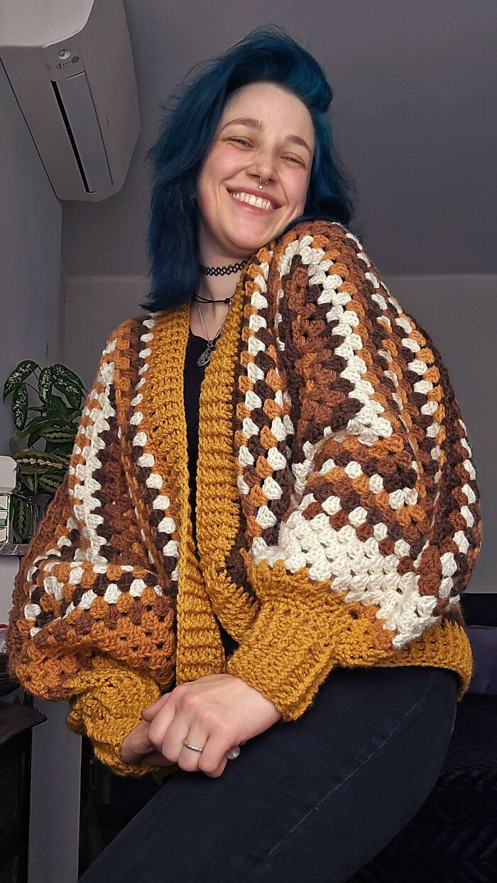 Smiling person in a colorful crocheted cardigan, featuring earthy tones, set in a cozy indoor environment.