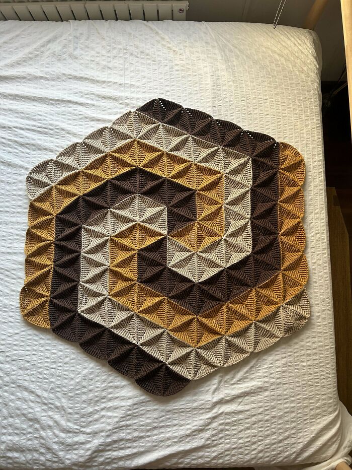 Hexagonal crocheted blanket with geometric pattern in brown, beige, and yellow tones on a white bedspread.