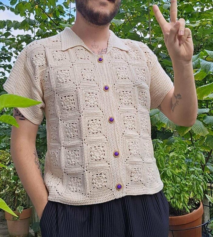 Man wearing crocheted shirt with purple buttons, standing outdoors, showing peace sign.