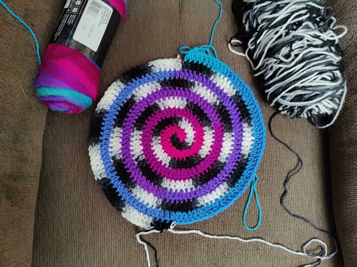 Crocheted spiral pattern in vibrant colors with yarn skeins nearby on a couch.