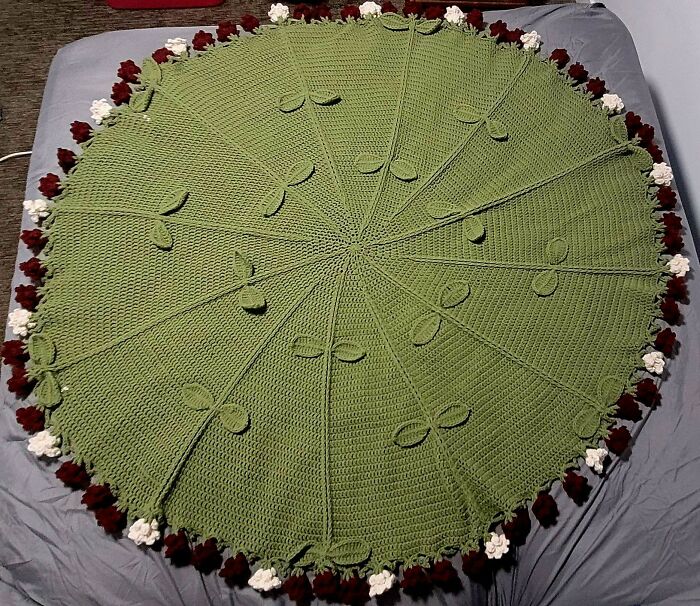 A crocheted green round blanket with red and white flower edges displayed on a bed.