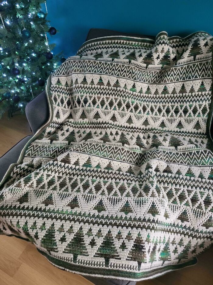 Crocheted blanket with intricate tree patterns draped over a chair next to a decorated Christmas tree.