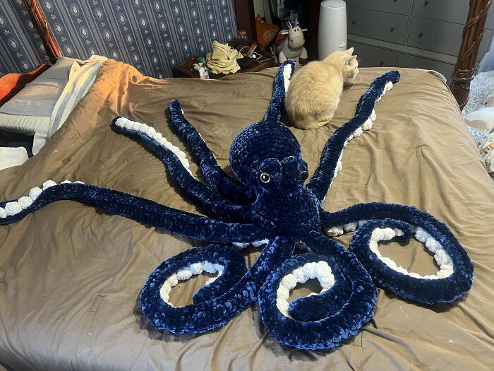 Crocheted blue octopus sprawled on a bed, with a cat sitting nearby.