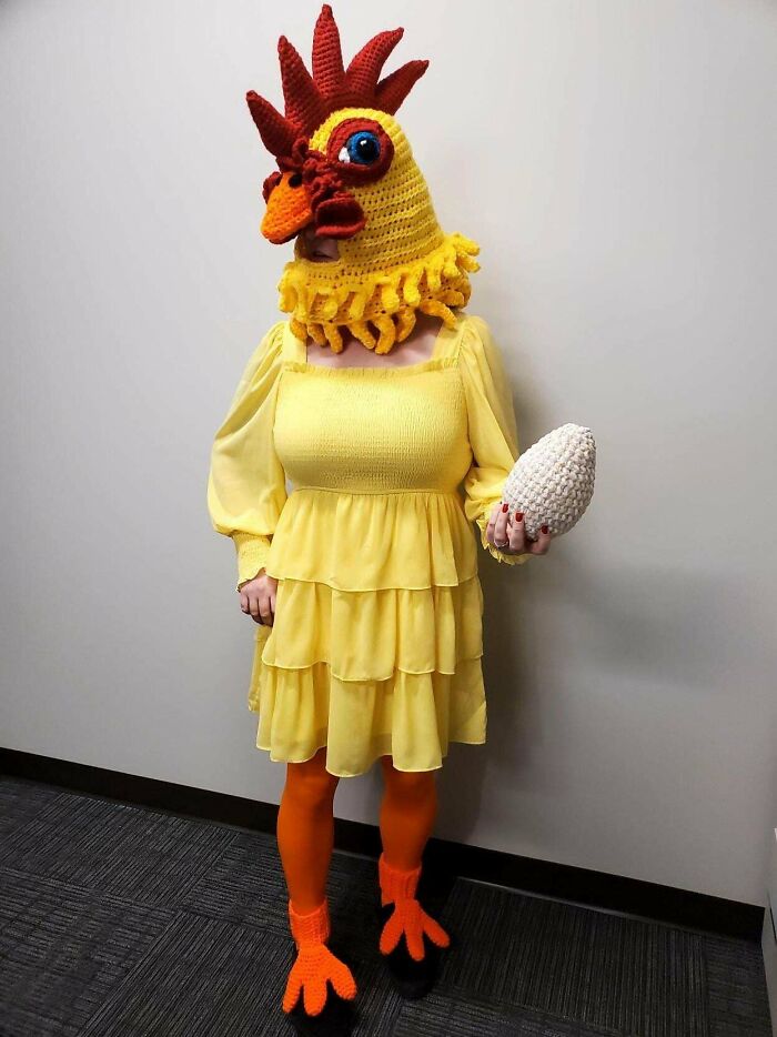 Person wearing a crocheted chicken costume, holding a crocheted egg, in a playful setting.