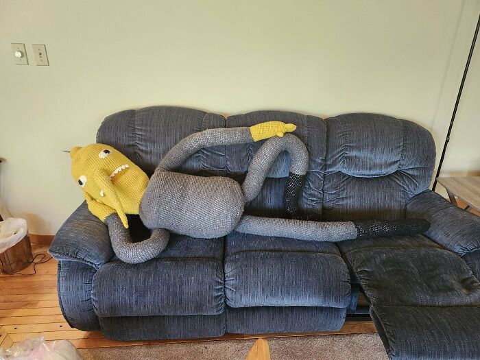 A crocheted yellow creature lounging humorously on a blue sofa with long limbs, showcasing creative crochet work.