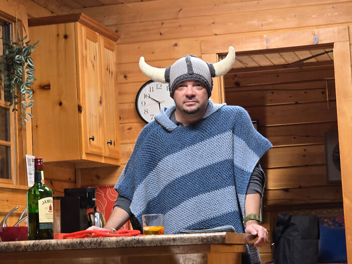 Man in a crochet Viking hat and blanket standing in a wooden kitchen.