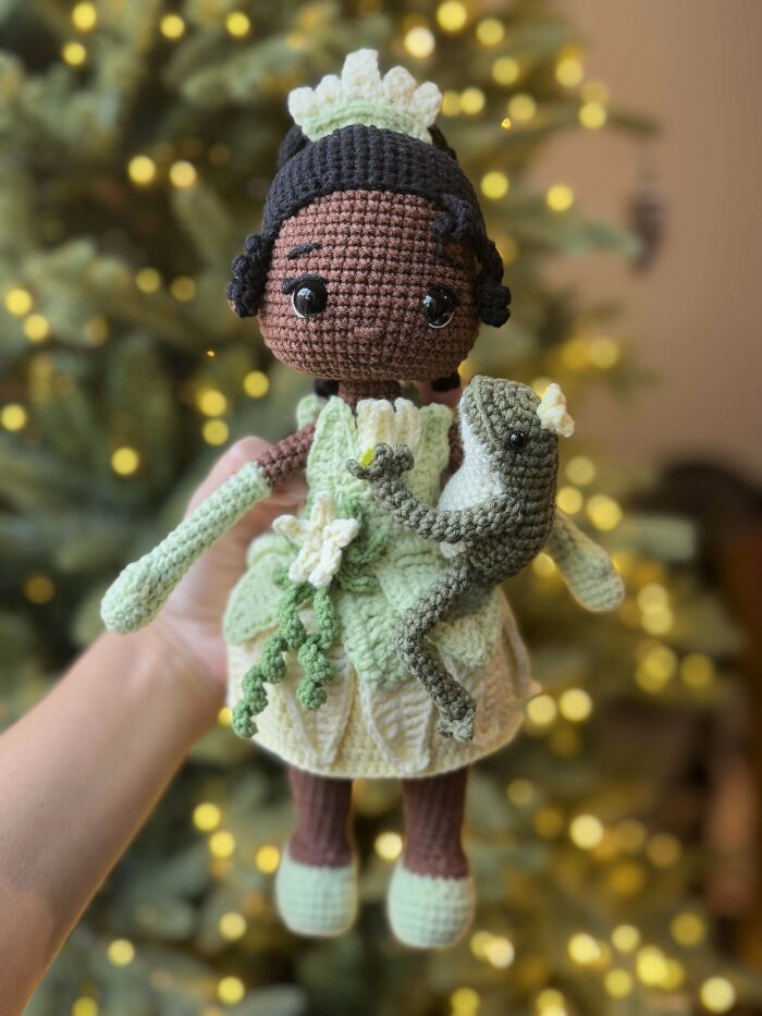 Crocheted doll in green dress holding a frog, with a blurred tree and lights in the background.