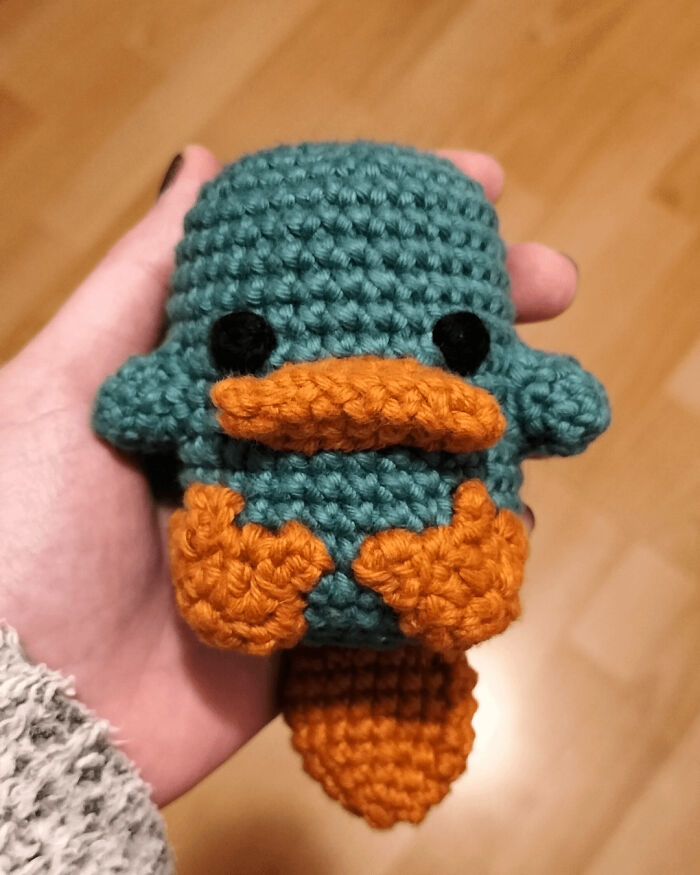 Crocheted plush platypus in teal and orange, held in a hand, showcasing creative craftsmanship.