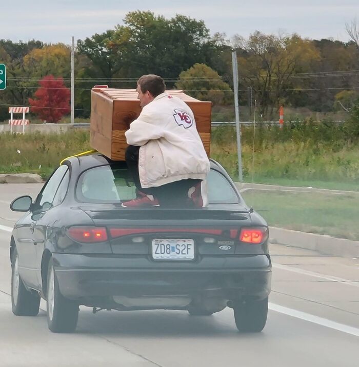 Person riding on top of a car holding a large wooden object, exemplifying risky behavior in traffic for Idiots-In-Cars theme.