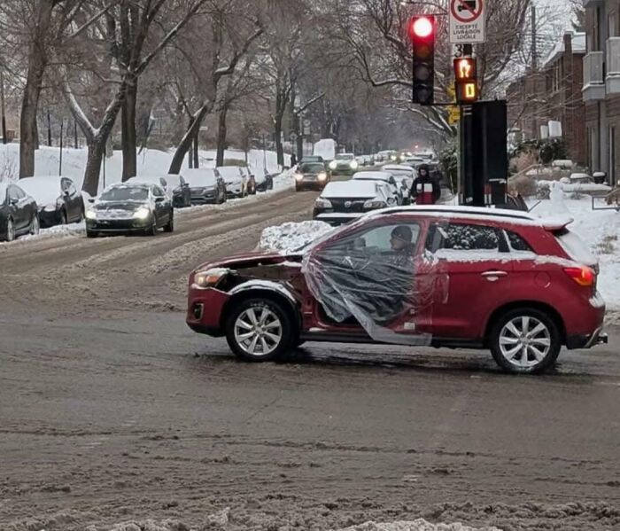 Red car with damage and plastic-covered door stopped at snowy intersection under red light.