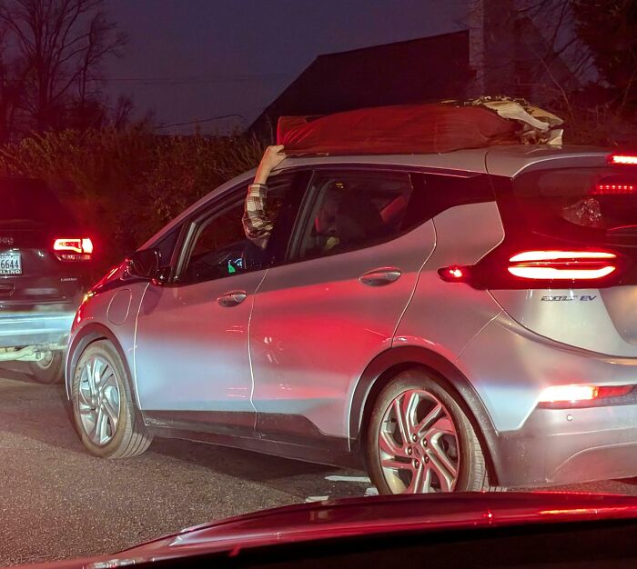 Person holding mattress on car roof in heavy traffic at night, illustrating risky behavior by drivers.