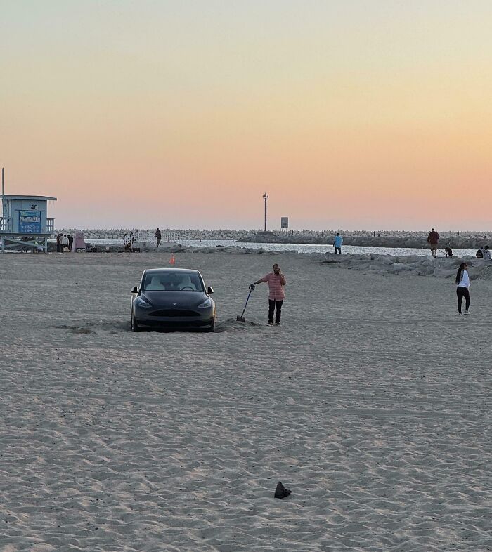 Car stuck on a sandy beach at sunset with a person digging near it.