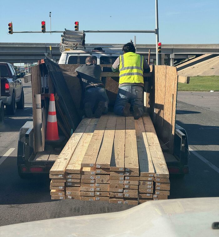 Two people kneeling on a trailer loaded with lumber at a stoplight, demonstrating dangerous behavior.