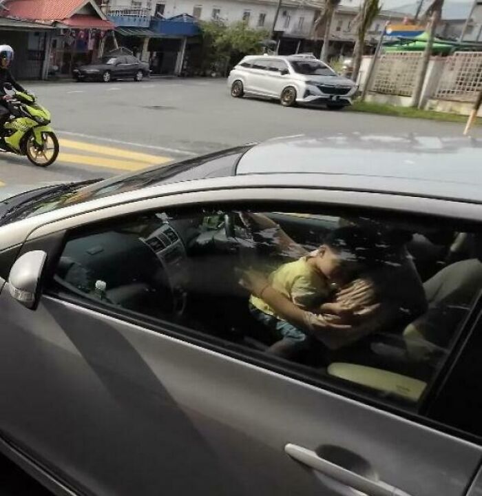 A child leaning on the steering wheel of a moving car, with another car and a motorcycle in the background.