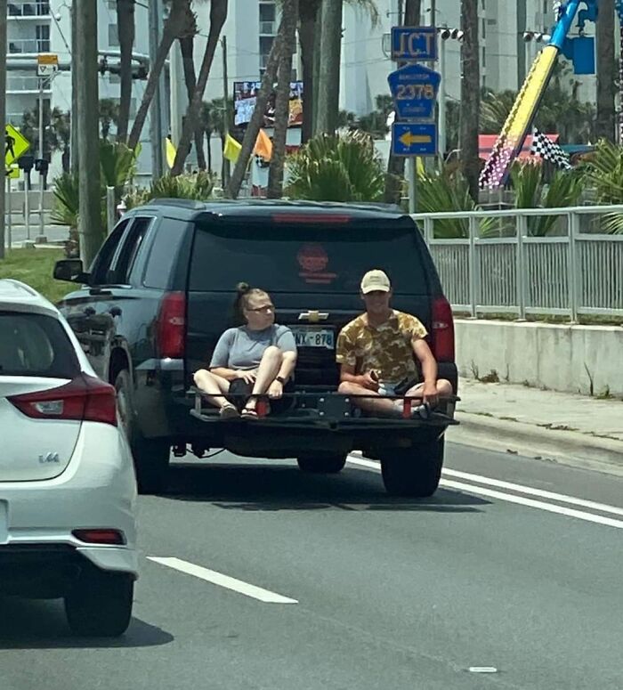 Two people sit dangerously on the back of a moving SUV on the road.