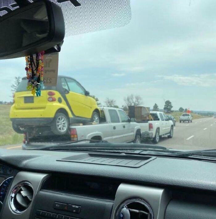 A yellow car precariously loaded onto a pickup truck bed on a highway, illustrating a classic 'idiots in cars' scenario.