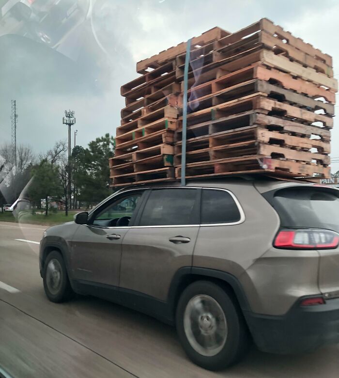 SUV driving with a precariously tall stack of wooden pallets on the roof, showcasing a typical "idiots in cars" scenario.