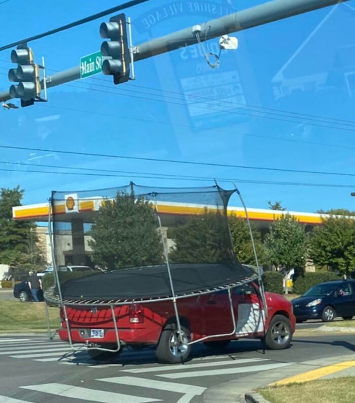 Red truck transporting a large trampoline unsafely, stopped at an intersection under clear blue skies.