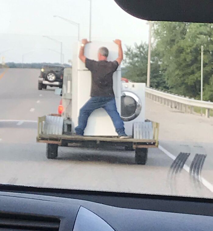 Man holding a washing machine on a trailer while traveling down a road, illustrating careless driving behavior.