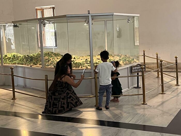 Adults and children examining a model landscape in a display case, with one adult using a phone.