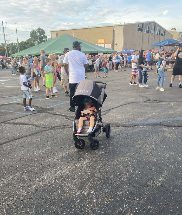 Child in a stroller using a mobile device at an outdoor event with crowd, highlighting distracted or ignorant-parents.