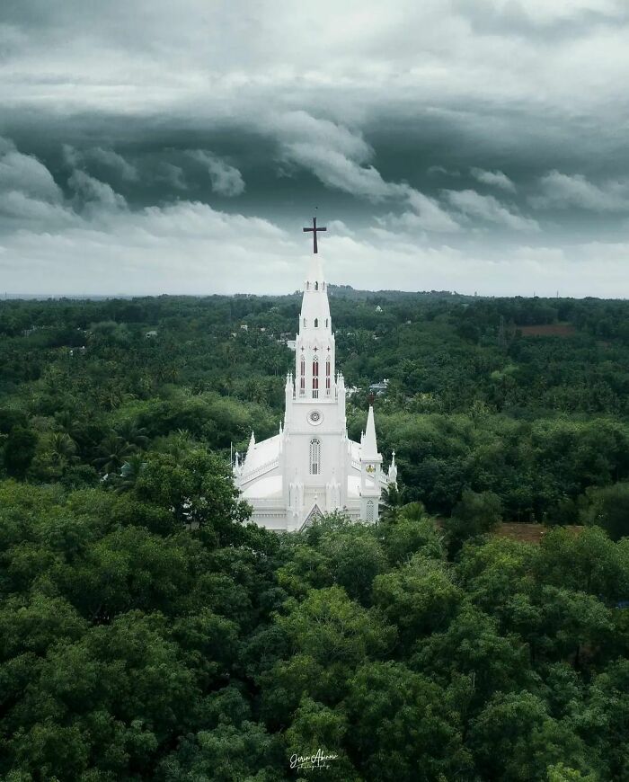 Aerial view of a Gothic-style church surrounded by lush greenery under dramatic cloudy skies.