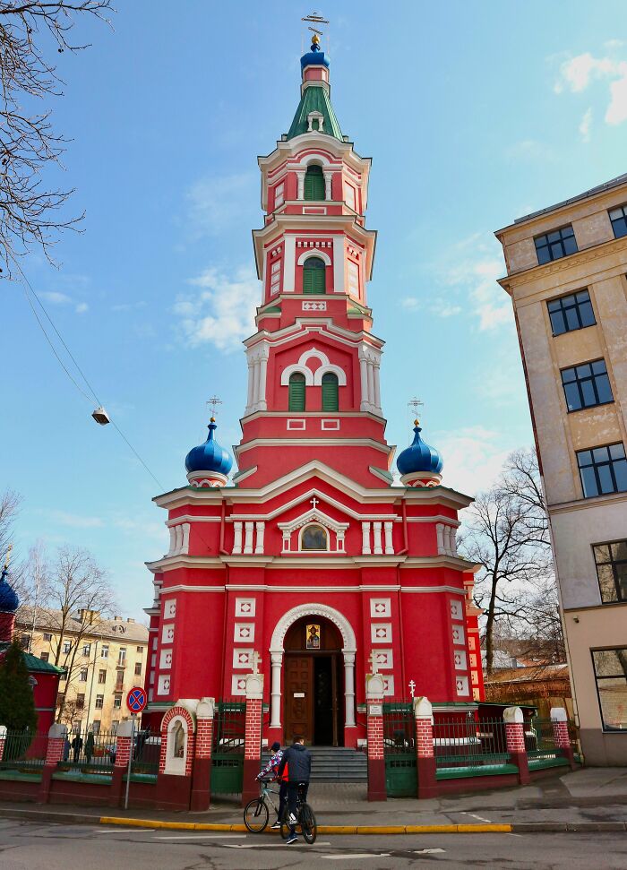 Red and white church with blue domes on a sunny day, featuring intricate architectural details and a cyclist in the foreground.