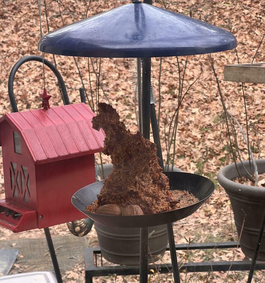 A bird feeder features a clump of seeds resembling a dinosaur shape, set against a background of fallen leaves.