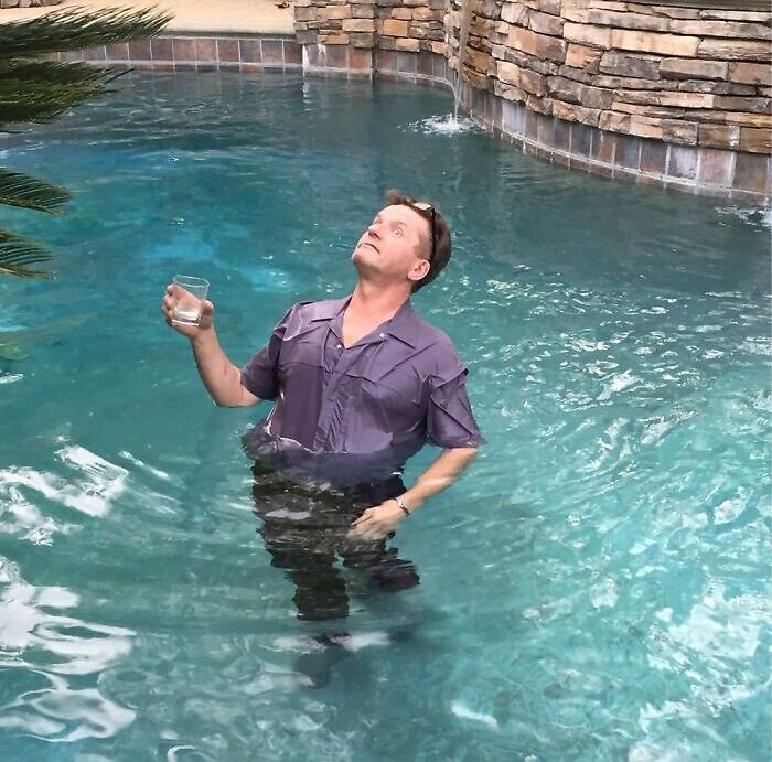 Man standing in a pool fully clothed, holding a drink, capturing a funny-camera-roll-find moment.